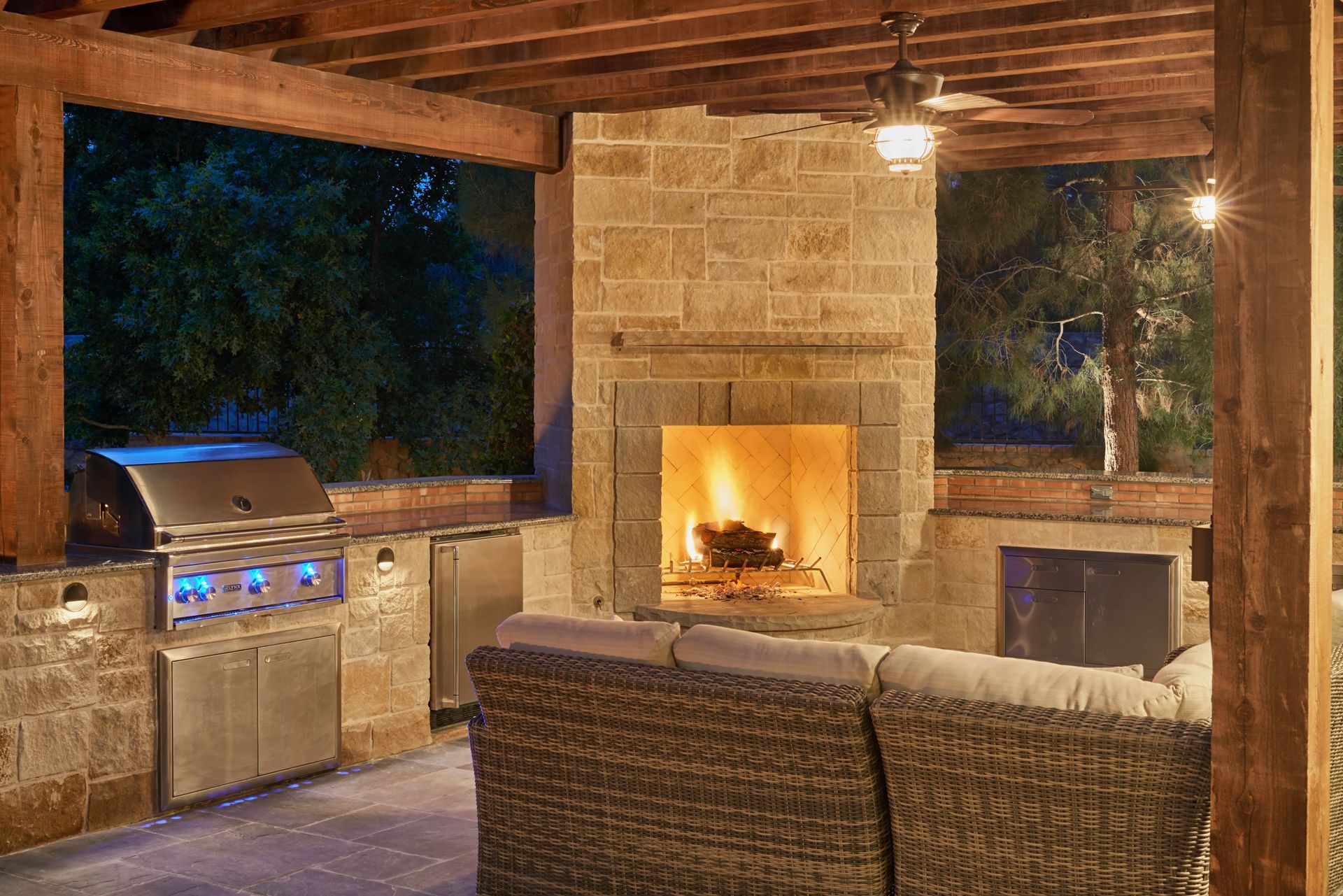 Outdoor stone fireplace and kitchen under a wooden pergola, illuminated at night.