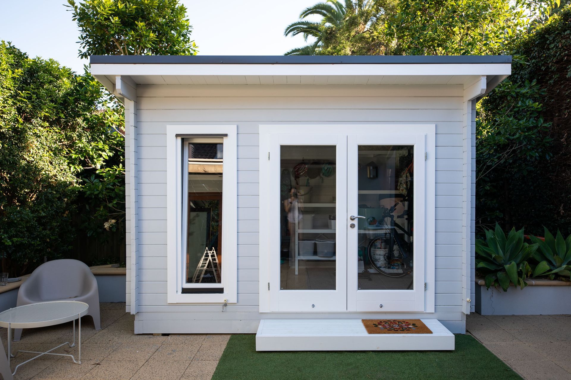 Light blue shed with white trim, double doors, and small window, set on patio with seating.