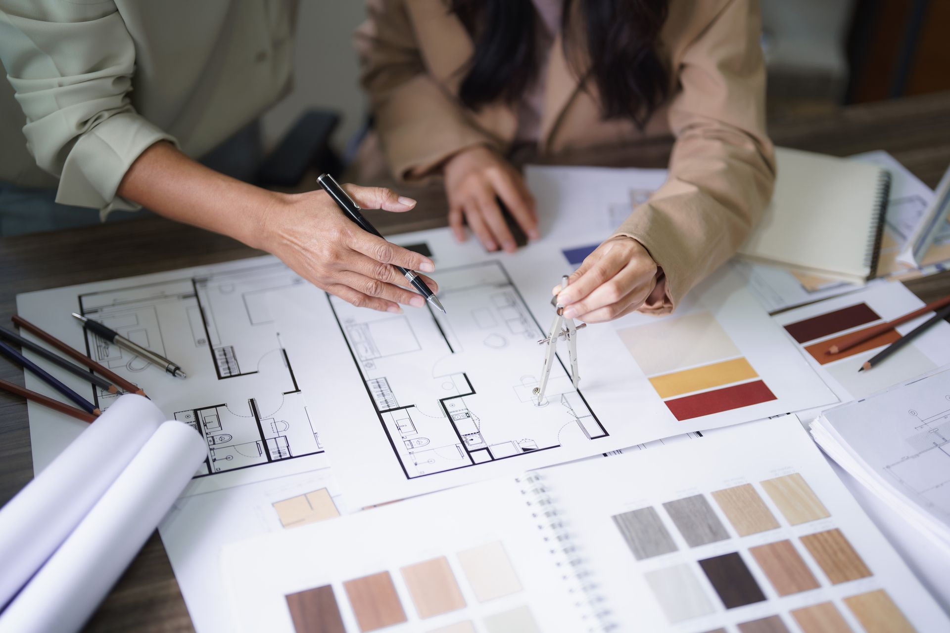 Two people reviewing architectural plans, using pens and compass, with color swatches on a desk.