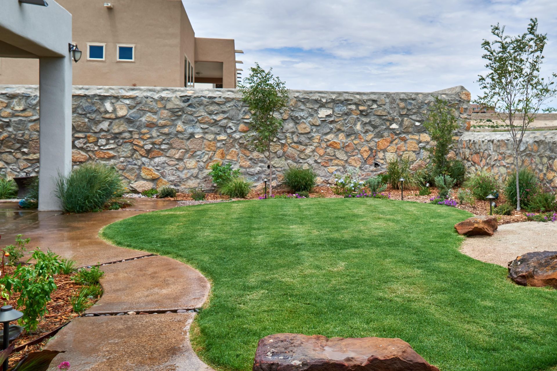 Lush green lawn in a backyard with stone wall and landscaping. Concrete path leads to the yard.
