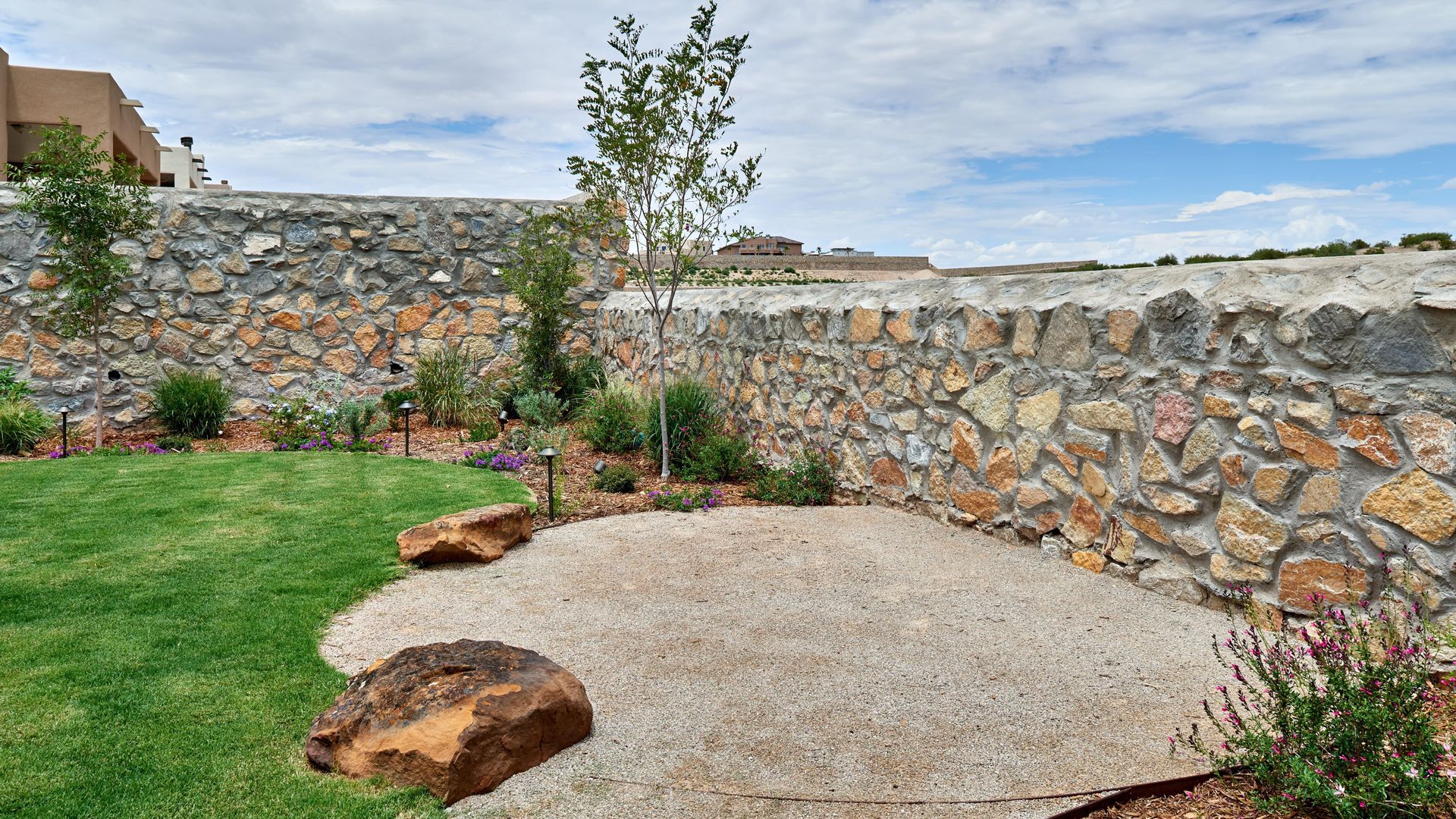 Stone wall surrounds a xeriscape garden with grass, gravel, and a small tree.