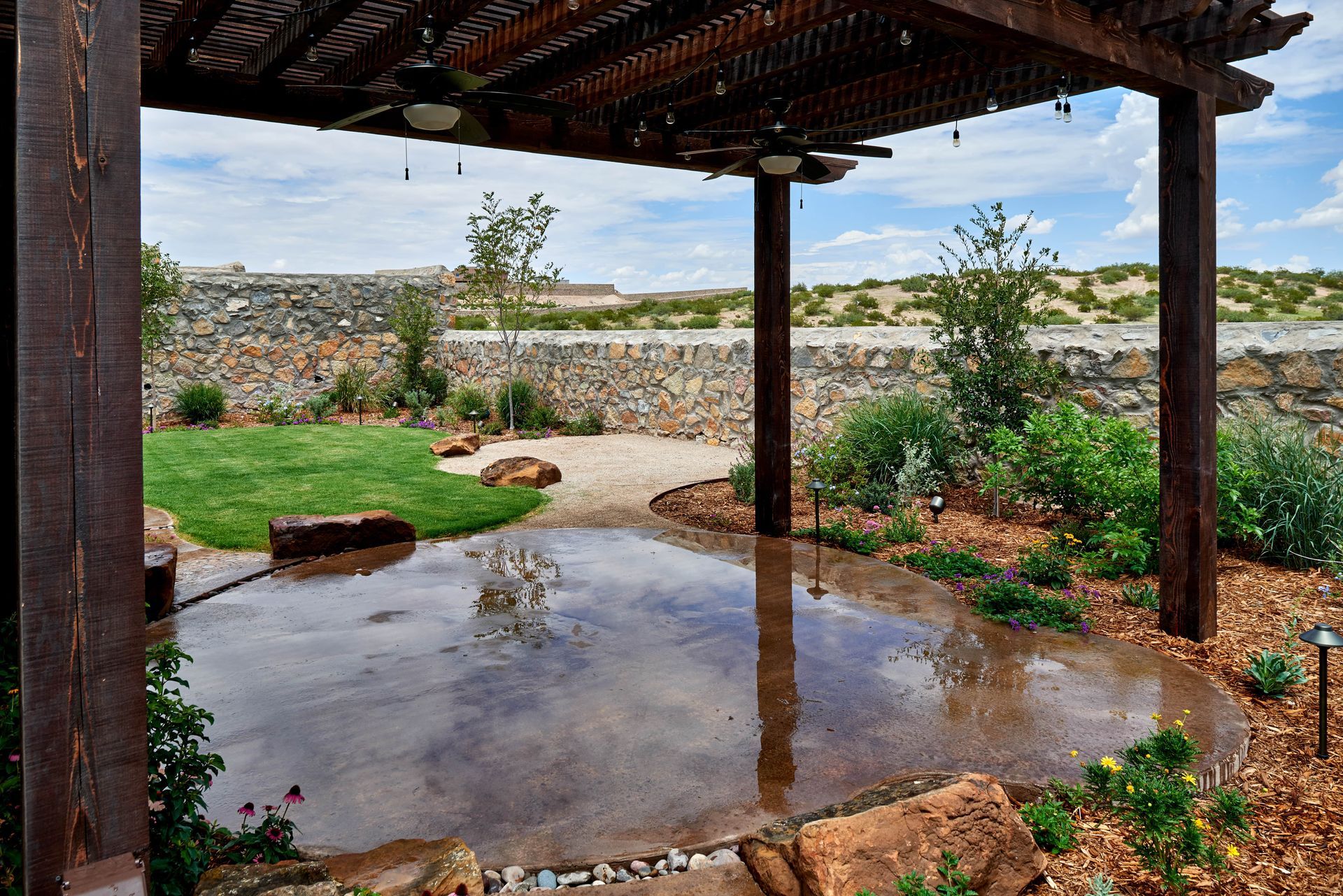 Pergola with concrete patio, grass, stone wall backdrop, and landscaping.