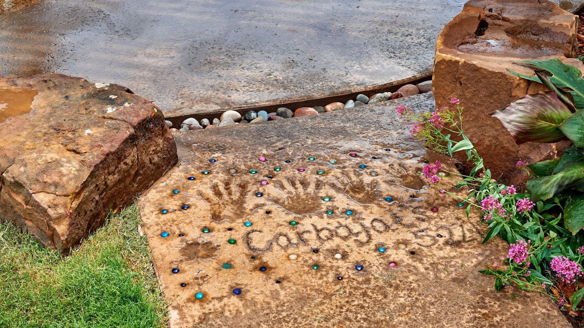Stone path with handprints and name “Carbajal 5-21”. Rocks frame the pathway, with green grass and flowers.