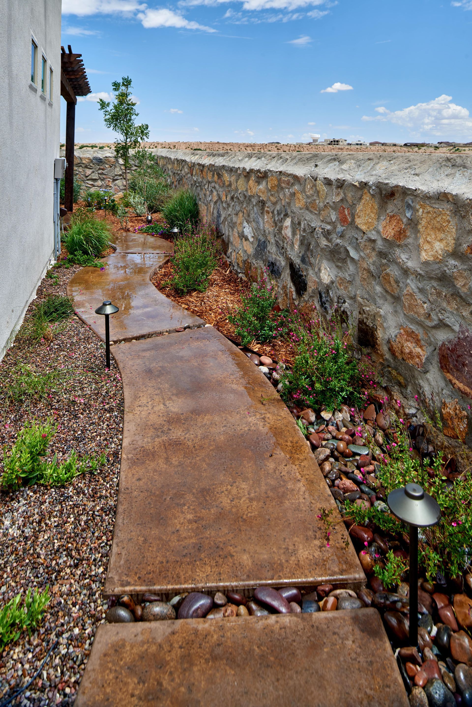 Concrete pathway winds through a garden with a stone wall and landscaping.