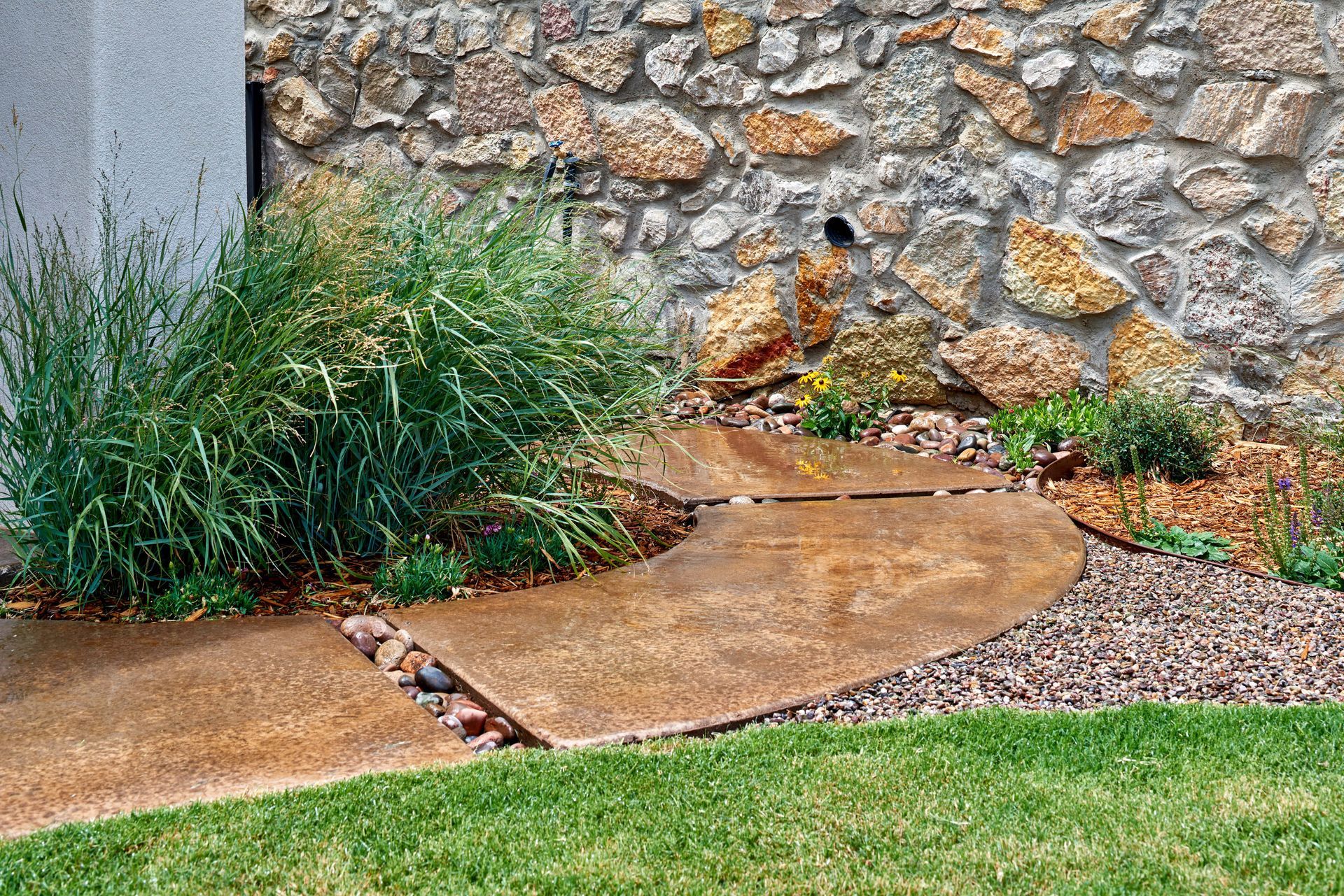 Curved rusty metal walkway through a garden bed with grass, rocks, and a stone wall.