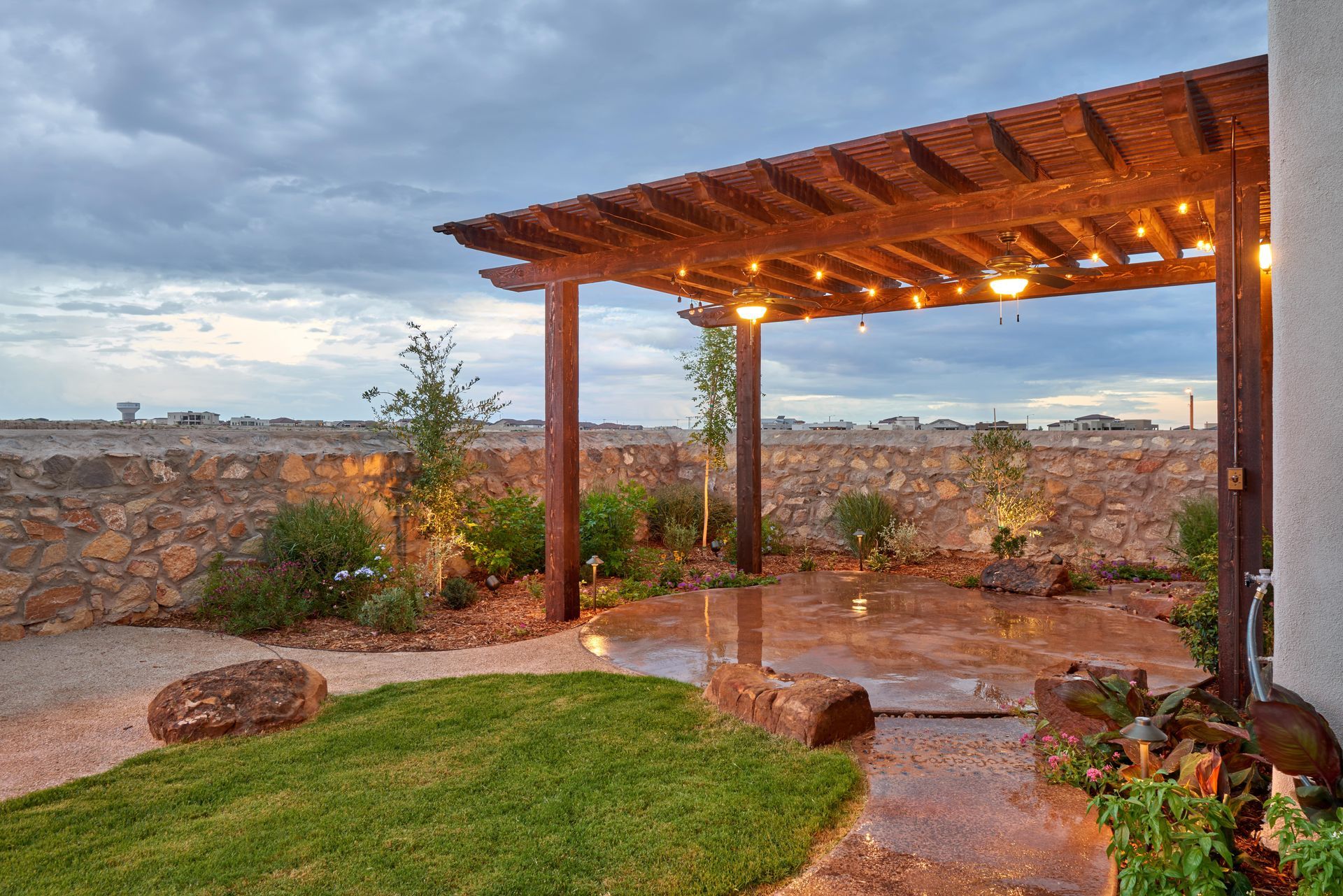 Patio with pergola strung with lights, stone wall backdrop, green lawn and plants, cloudy sky.