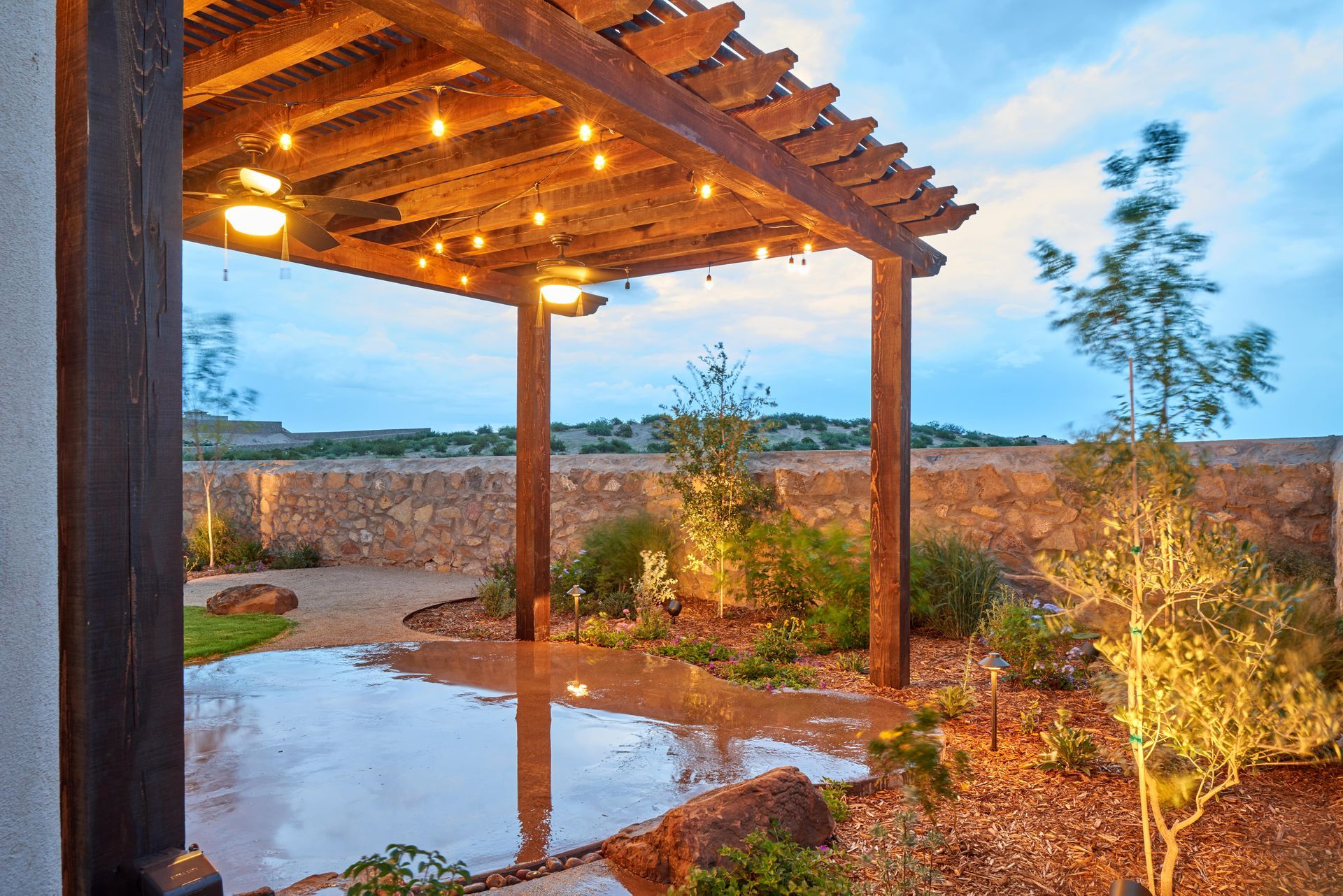Pergola with string lights over a patio with reflecting water, surrounded by plants and a stone wall.
