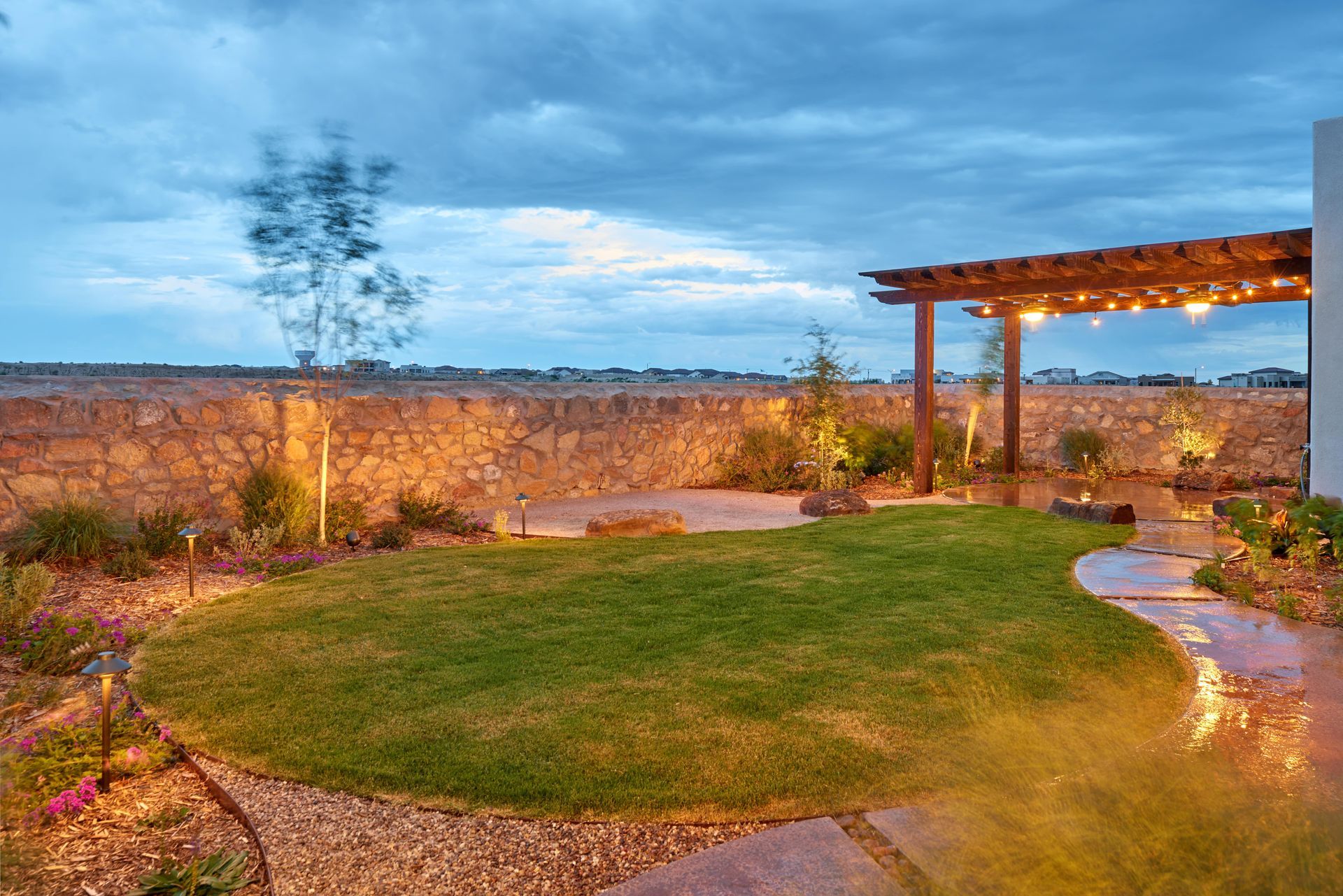 Backyard with green grass, stone wall, wooden pergola, and string lights under a cloudy sky.