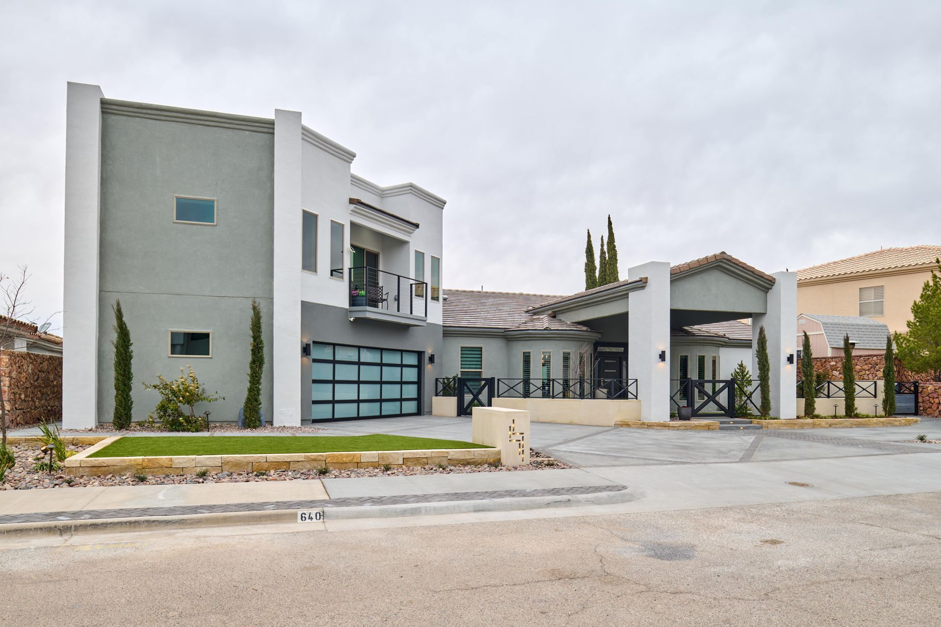 Modern two-story house with gray and white stucco exterior, glass garage door, and a covered entryway; a cloudy sky overhead.