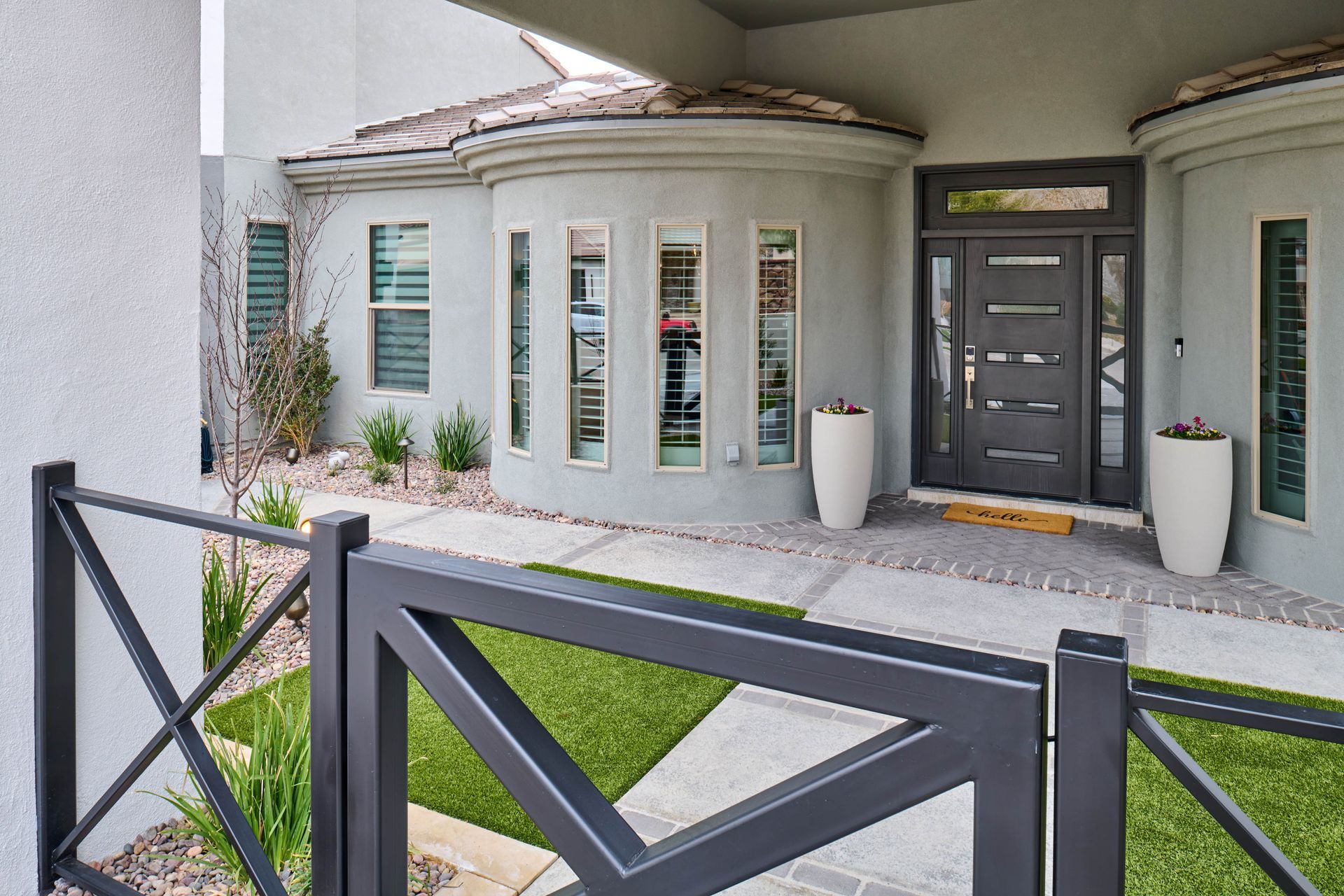 Grey modern home entrance with dark door, windows, and decorative planters. Black gate in foreground.