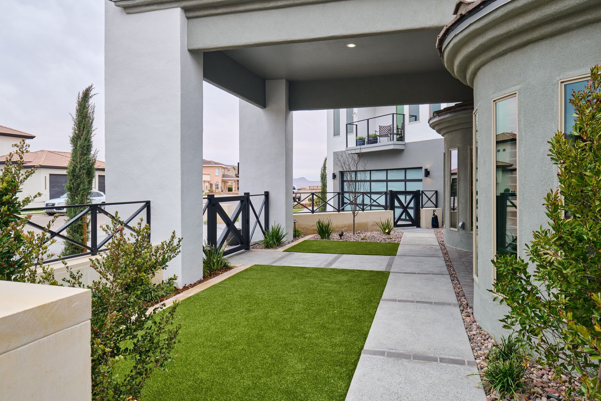 Pathway with artificial turf, bordered by shrubs and a black gate, leads to a modern building.