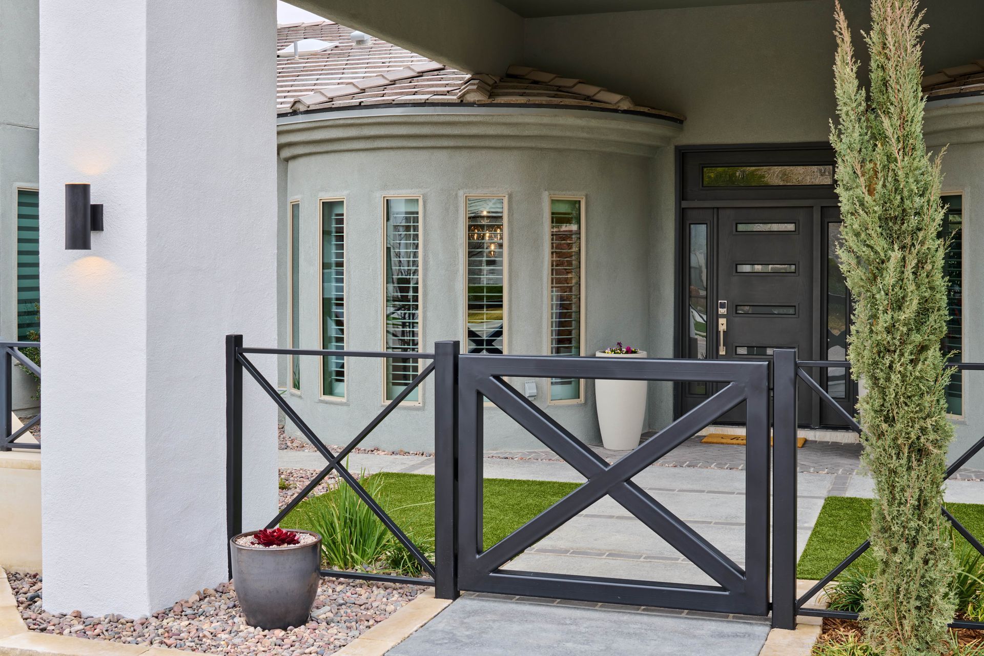 A dark gray front gate with an X-shaped design, leading to a house with a curved front.