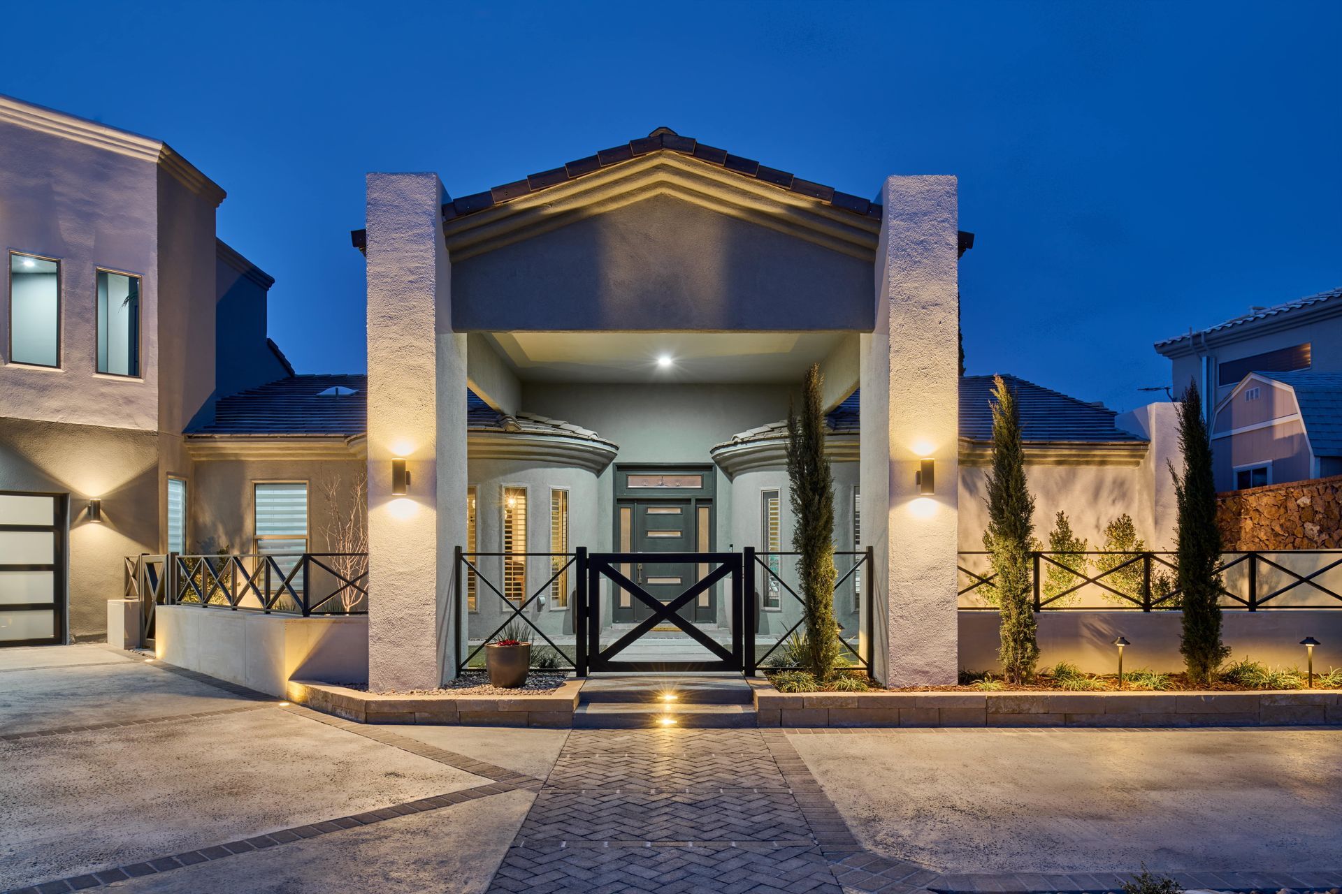 Front exterior of a modern house at night with illuminated pillars and a black gate.
