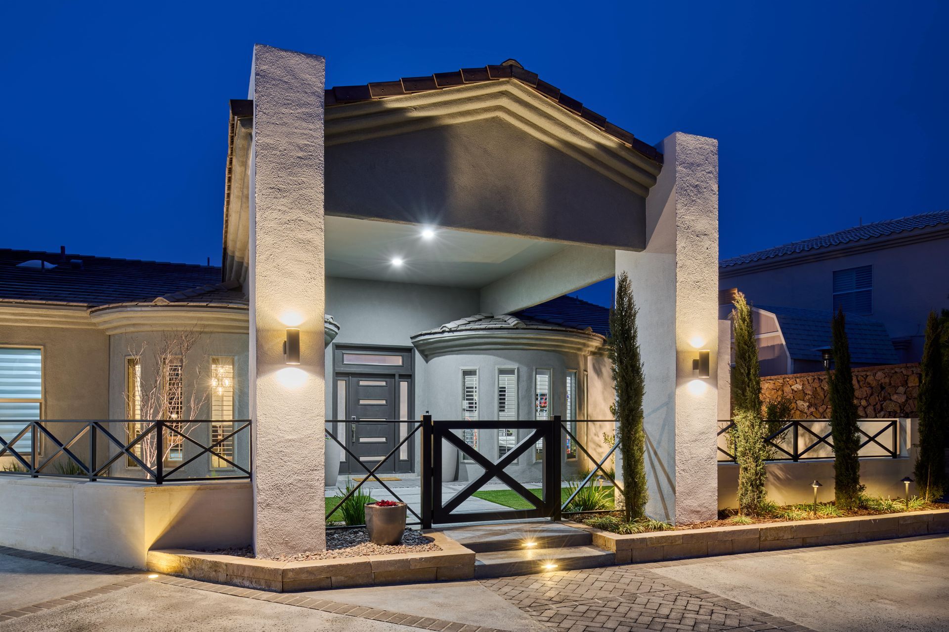 Night view of a modern home with illuminated pillars, entry gate, and landscaping against a dark blue sky.