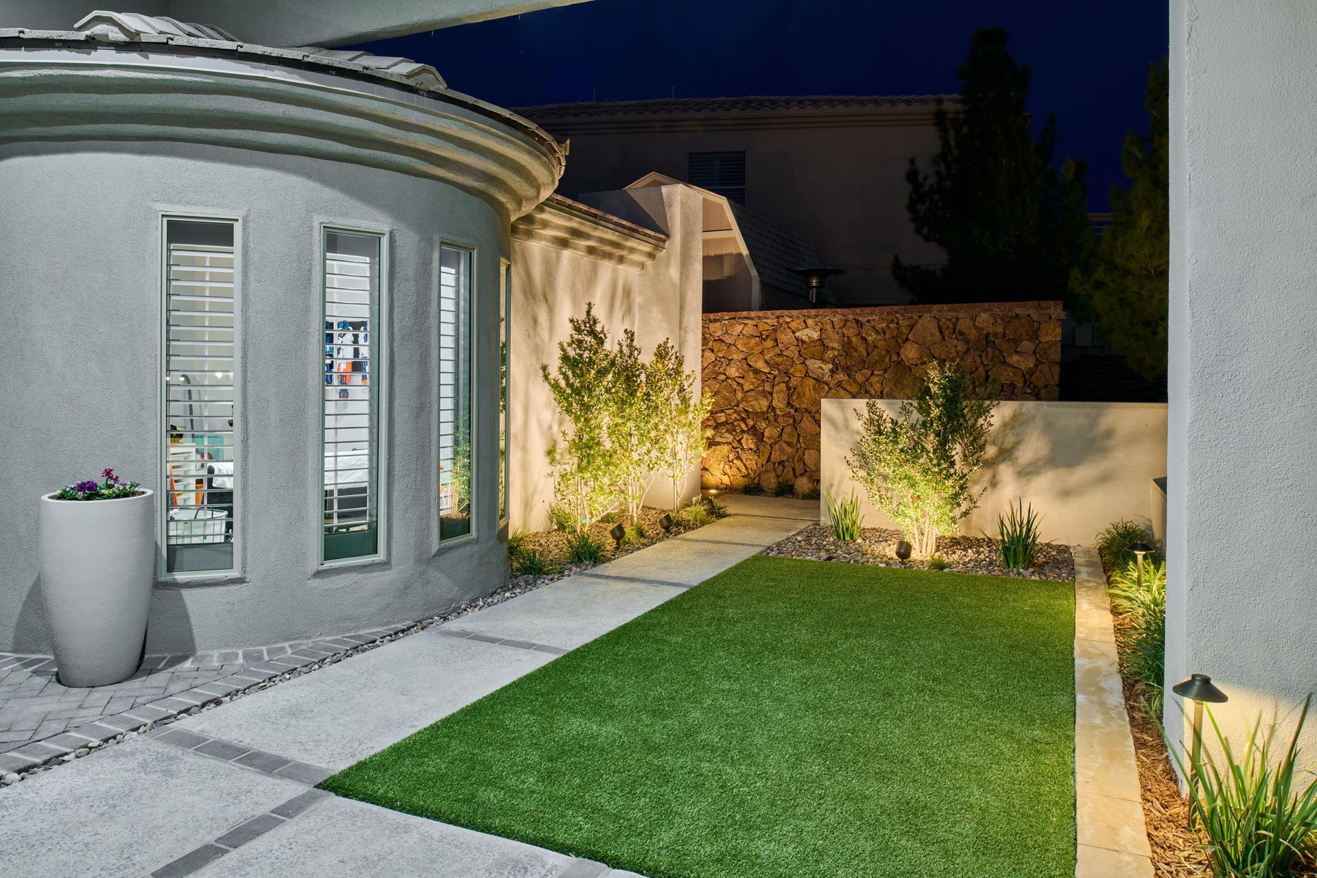 Night view of a modern home exterior with lighted pathway, green lawn, and stone wall backdrop.