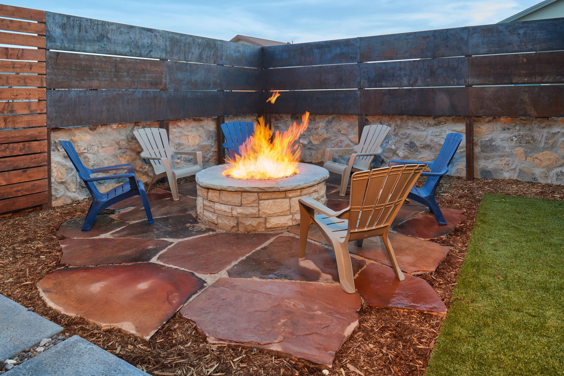 Fire pit area with stone pavers, Adirondack chairs, and a metal fence.