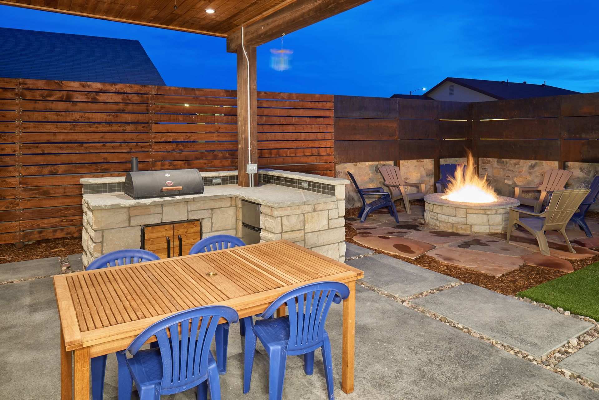 Outdoor patio with grill, fire pit, dining table, and blue chairs. Wooden fence and overhead beam. Nighttime setting.