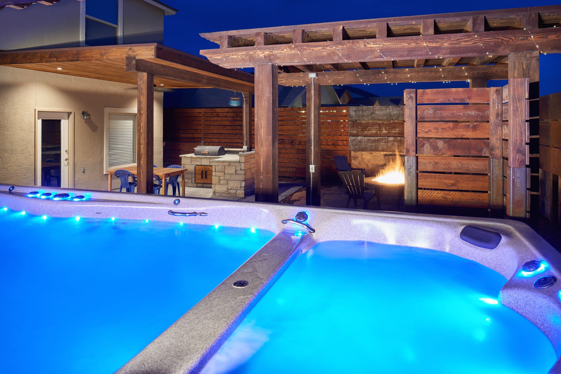 A backyard hot tub and pool illuminated with blue lights at night, next to a wooden pergola and outdoor kitchen.
