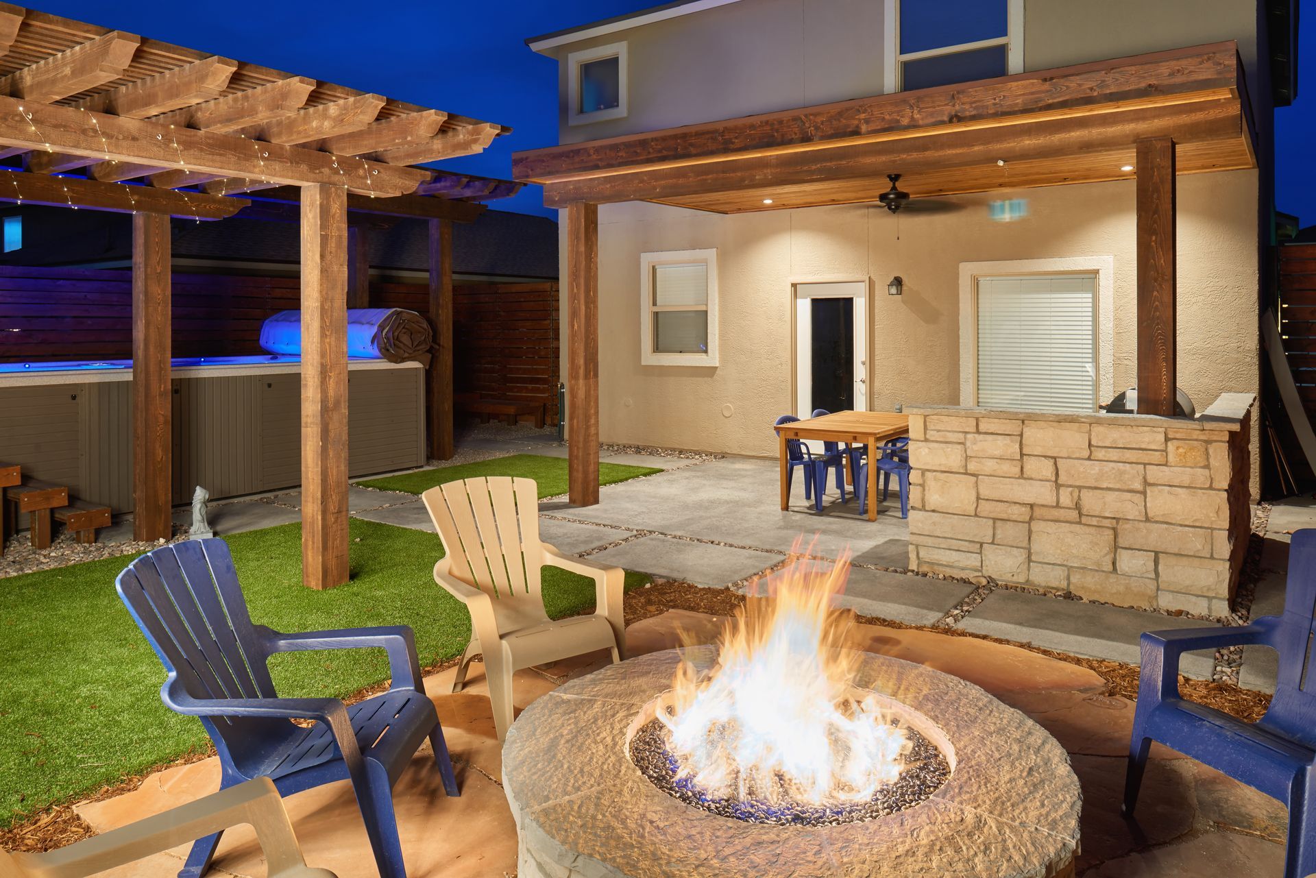 Backyard patio with fire pit, hot tub, seating, pergola, and house at night.