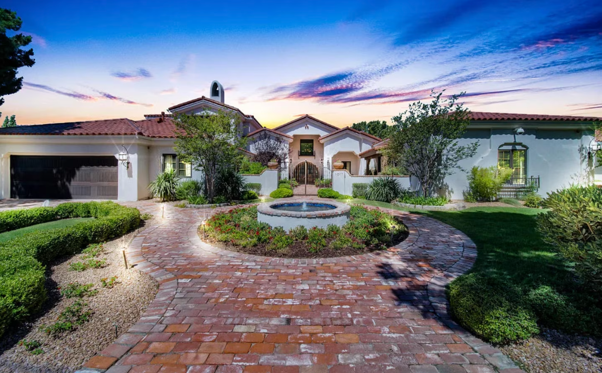 Elegant home with red tile roof, brick path, fountain, and manicured landscaping under a sunset sky.