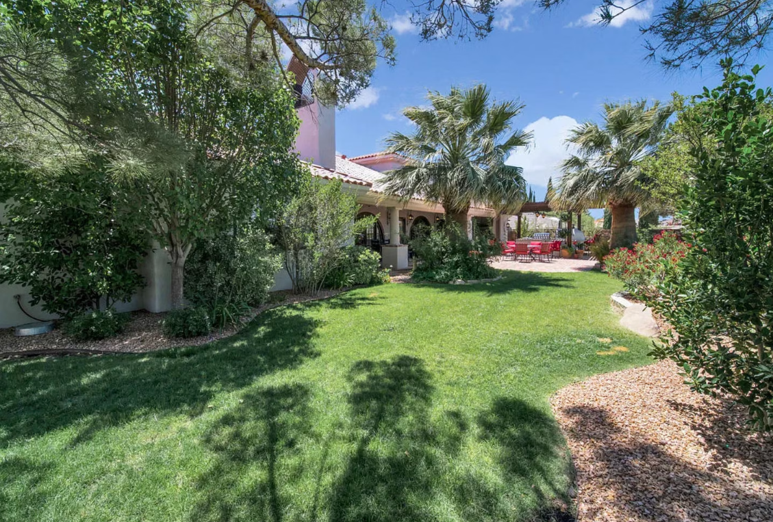 Green lawn with trees and shrubs in a sunny backyard, red patio furniture in the distance.