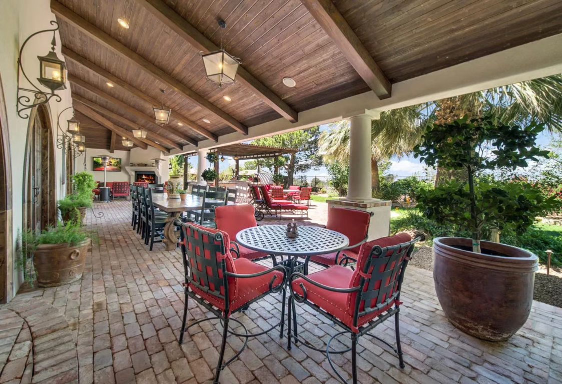 Covered patio with red seating, dining table, and brick flooring, overlooking greenery.