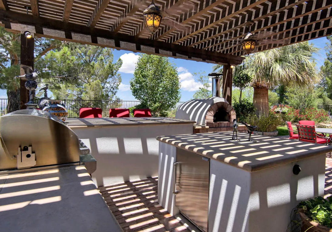 Outdoor kitchen with pergola, pizza oven, and seating; sunny day with trees in the background.