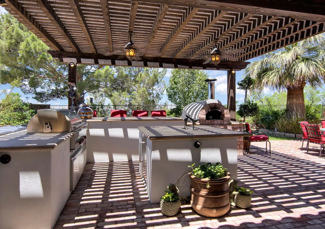 Outdoor kitchen with grill, pizza oven, and pergola, on a sunny patio.
