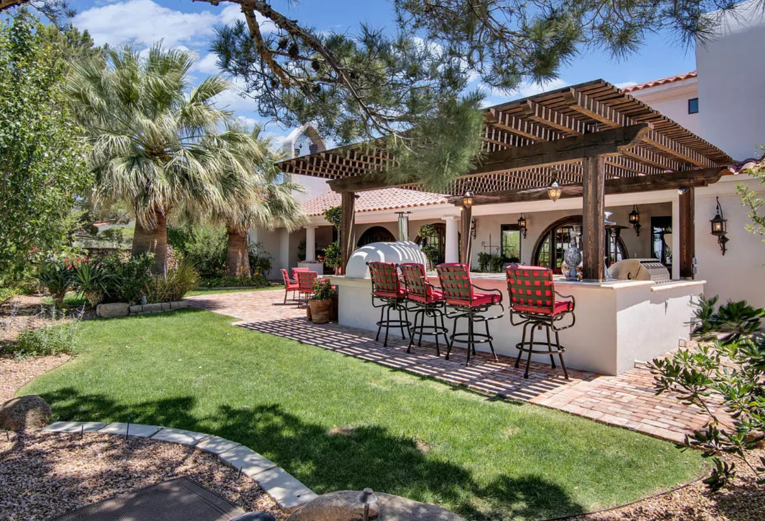 Outdoor patio with bar seating under a wooden pergola, adjacent to a house and grassy yard.