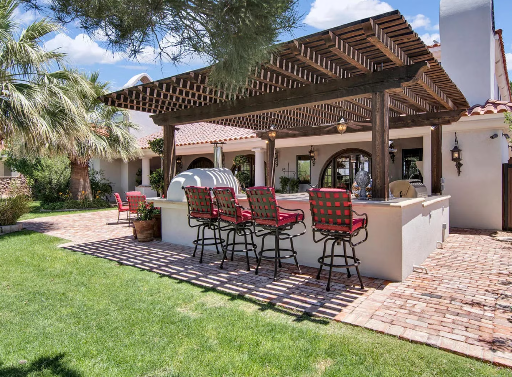 Outdoor kitchen with a pergola, bar, and seating on a patio, surrounded by grass and palm trees.