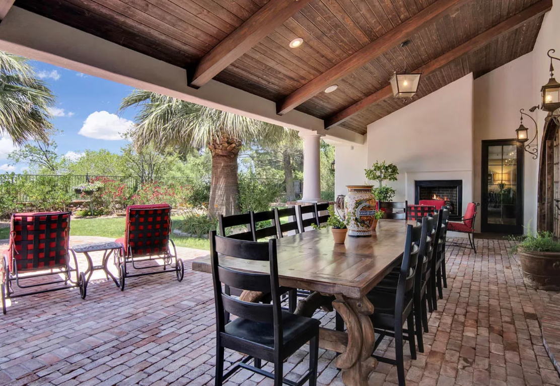 Covered outdoor dining area with a long wooden table, chairs, fireplace, and red brick floor.