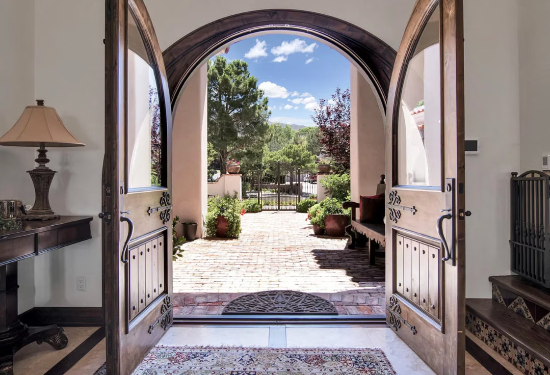 Open wooden doors frame a brick path leading to an outdoor courtyard with trees and a blue sky.