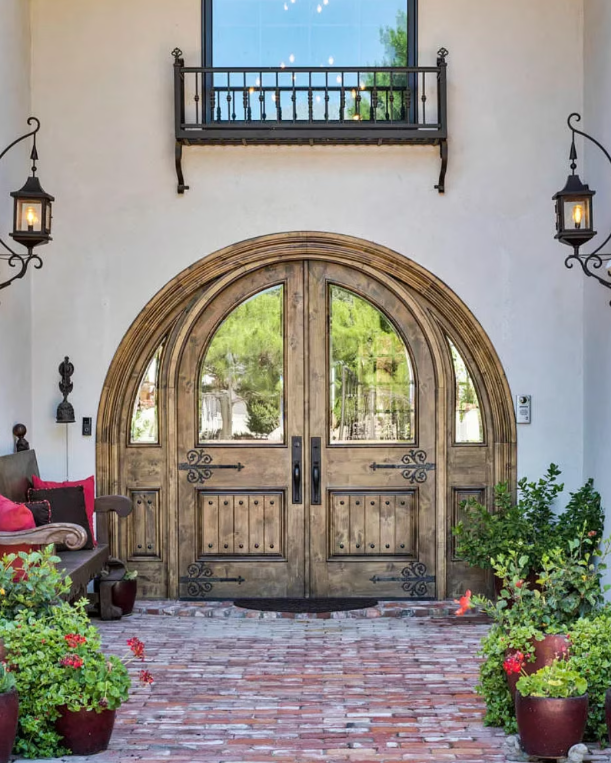 Arched wooden double doors with glass panels, flanked by lanterns and potted plants on a brick pathway.