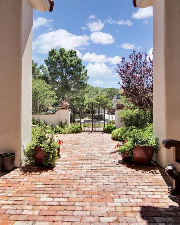 Brick pathway leads through arched entryway to a garden gate under a bright blue sky.
