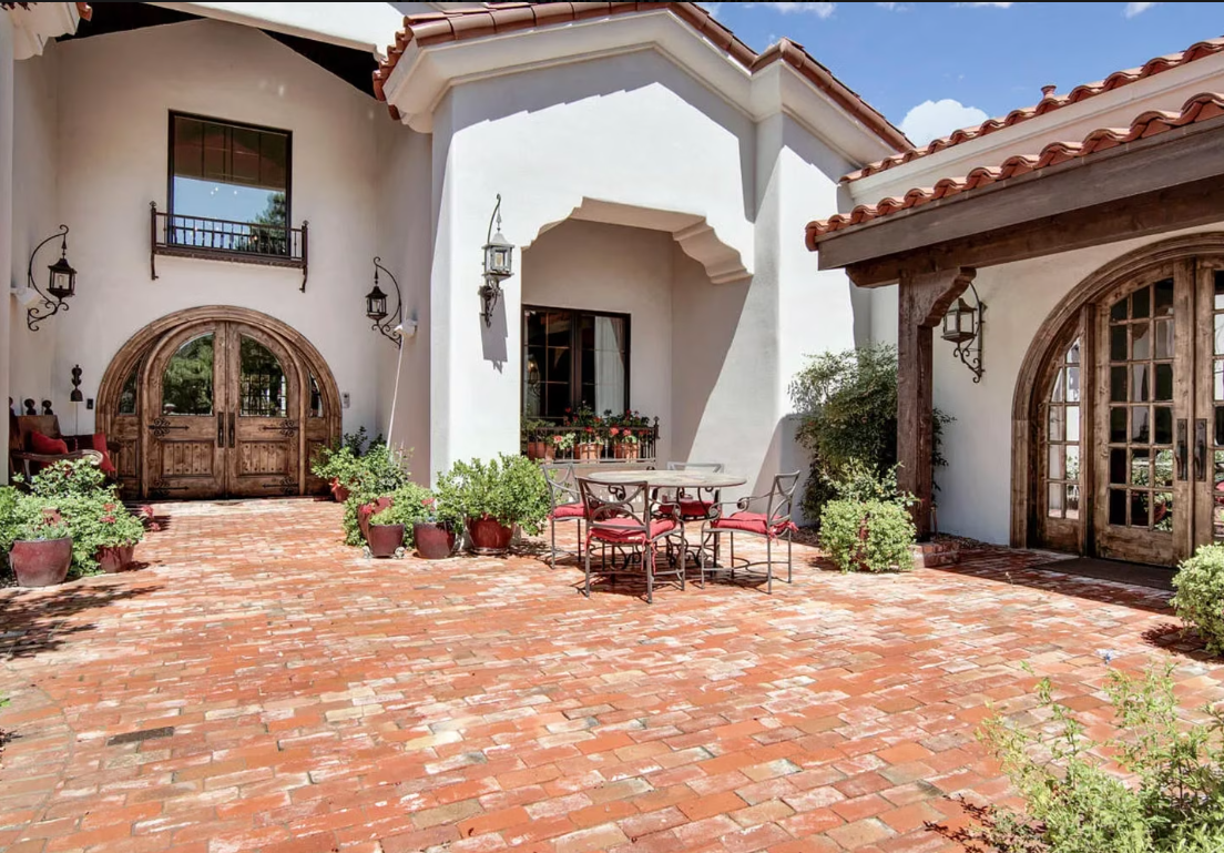Courtyard with brick pavers, white stucco building, arched doorways, red tile roof, and potted plants.