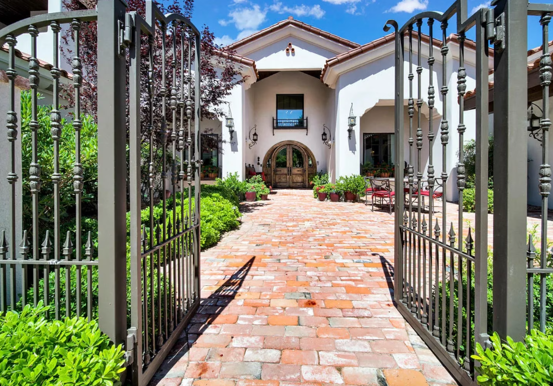Open wrought-iron gates leading to a white stucco house with a red-brick pathway and lush greenery.