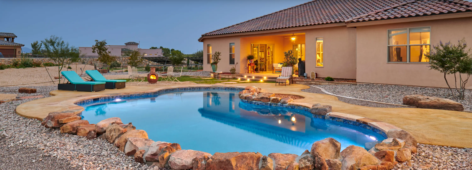 A pool with rocky border in front of a house. Two chairs sit near the pool, and the house has lights on.