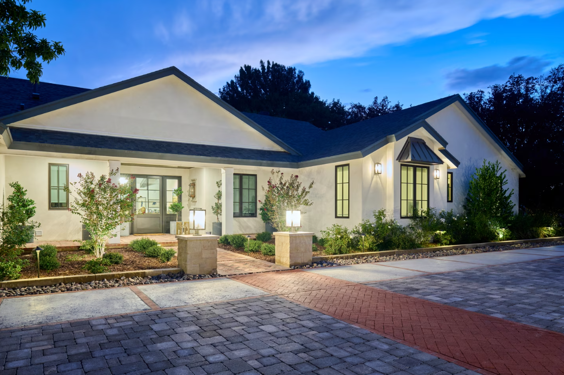 White stucco house with dark trim, brick walkway, and landscape lighting at dusk.