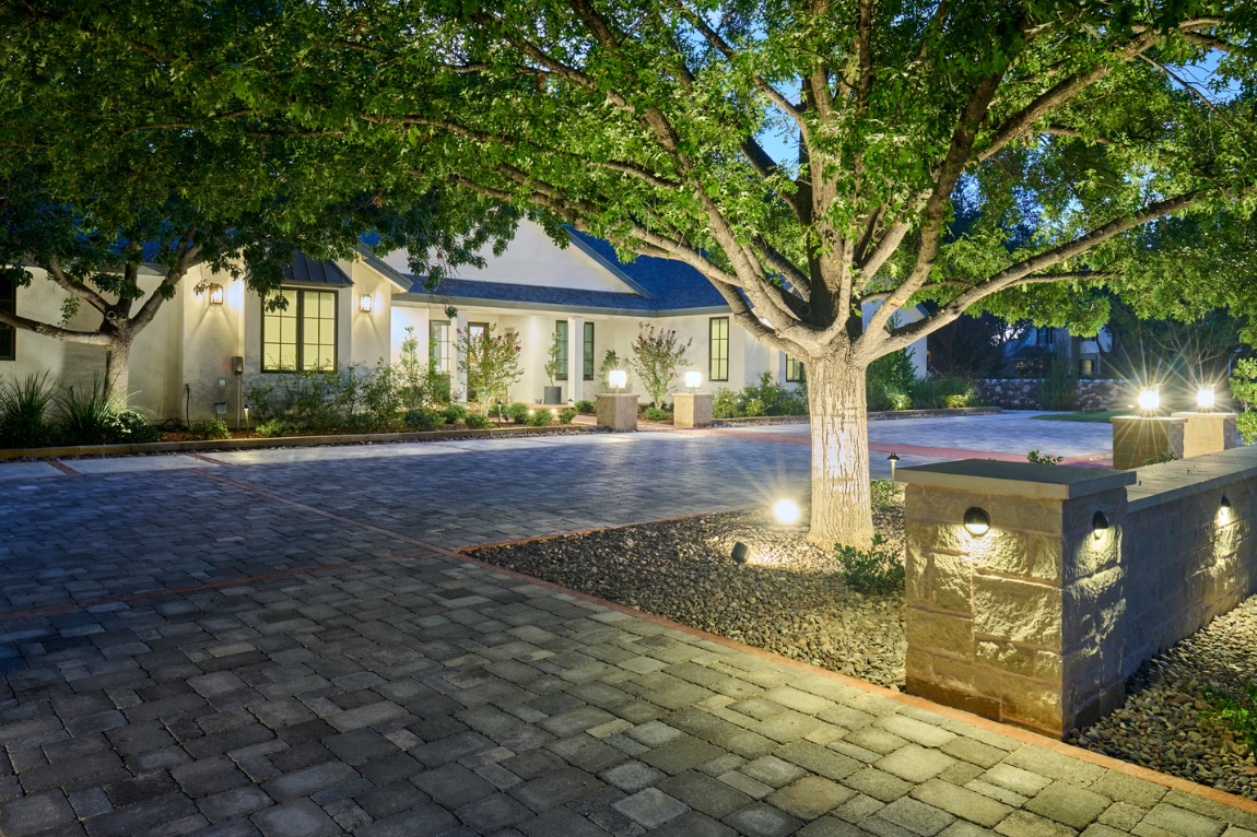 Cobblestone driveway leading to a white house, illuminated by outdoor lights under a large tree.