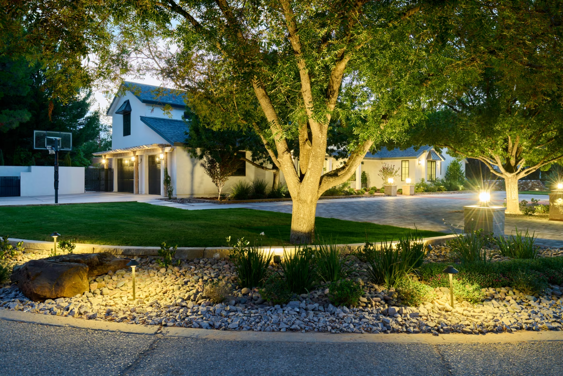 A house with outdoor lighting, a basketball hoop, and trees at dusk.