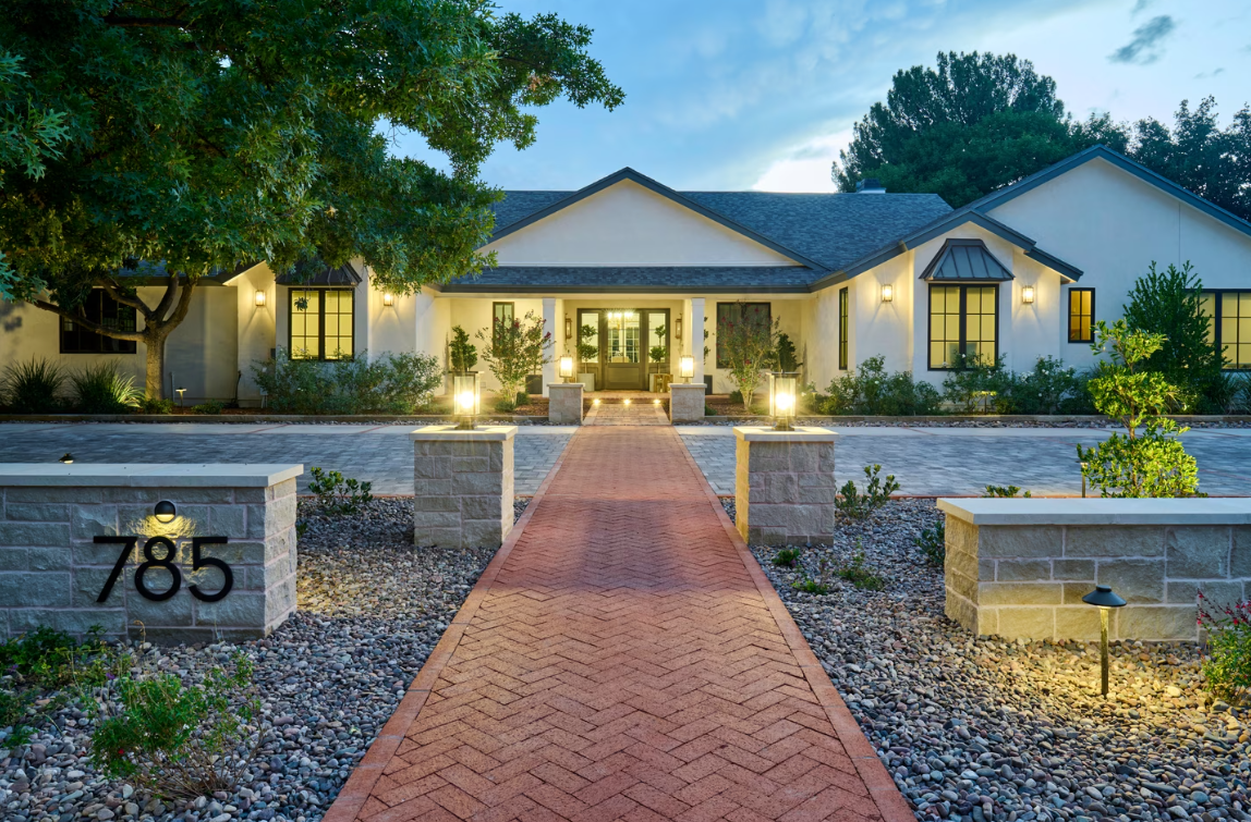 Elegant home with a brick path leading to the front door. Lit columns flank the path; gravel and plants in the foreground.