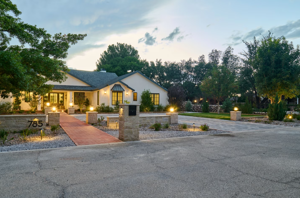 A well-lit house at dusk, featuring a brick walkway, stone pillars with lights, and a gravel driveway.