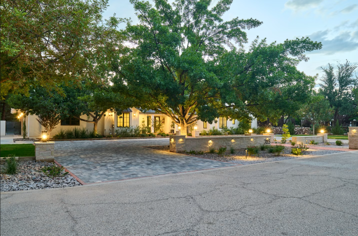 Stone-paved driveway leads to a white house, surrounded by trees. Illuminated low walls border the driveway.