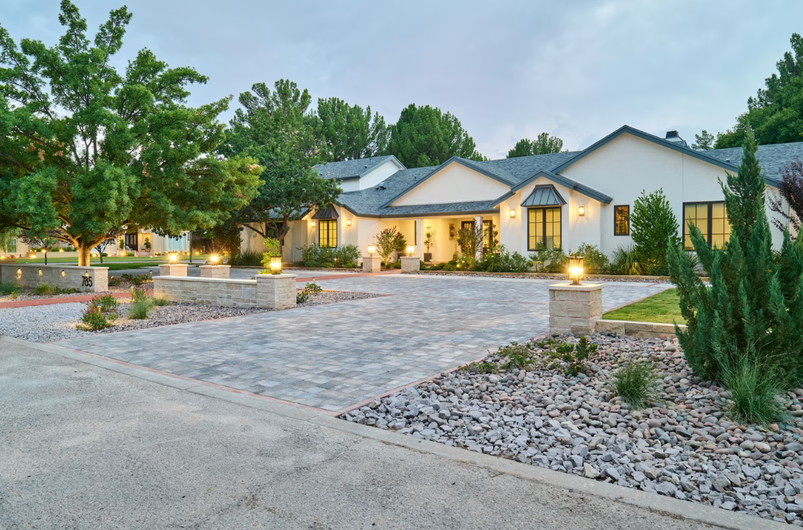 White house with a gray paved driveway, surrounded by trees and landscaping.