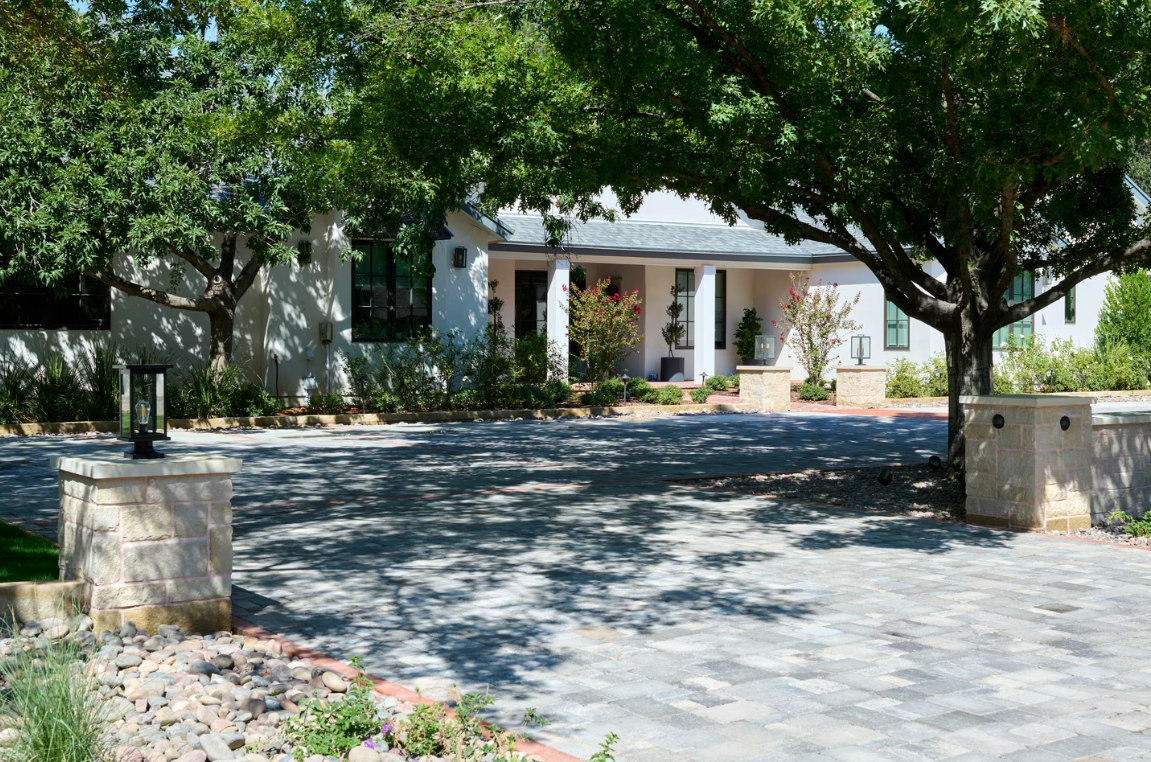 A white house with a stone driveway, flanked by trees and stone pillars.