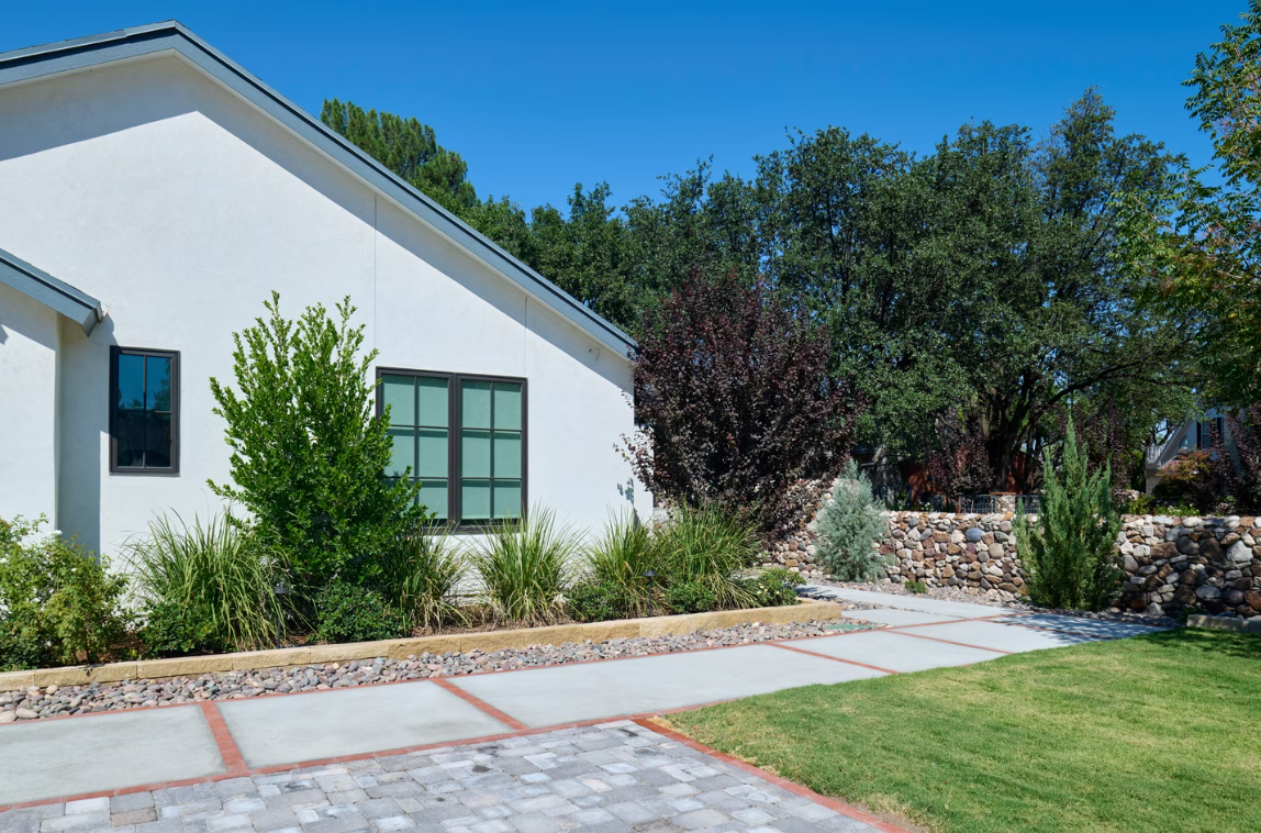 White house exterior with landscaping, stone wall, concrete path, and green lawn under a blue sky.