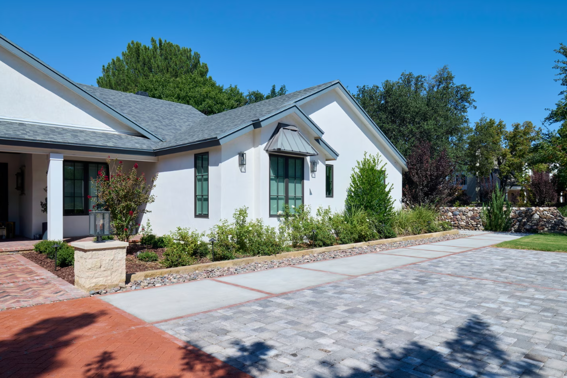 White house with blue sky, gray roof, and stone driveway, surrounded by greenery.