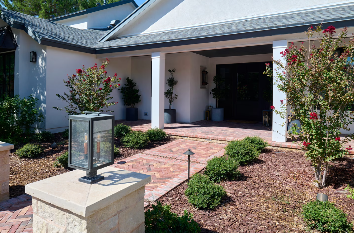 Brick pathway leads to a white stucco house with a covered entrance, lined with bushes and flowers.