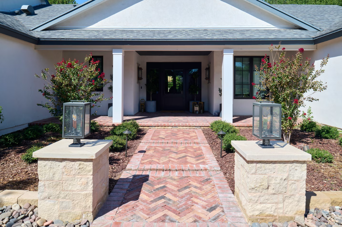 Brick pathway leading to a white house with pillars, flanked by lanterns and shrubs.