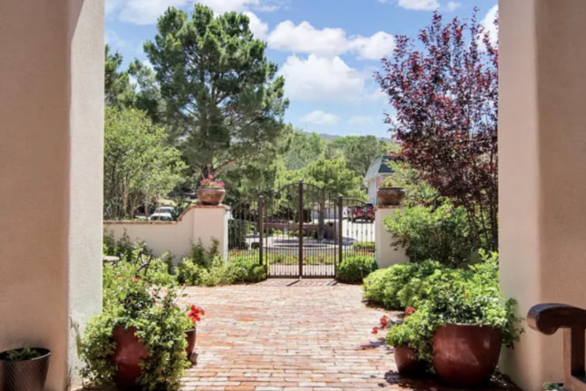 Brick pathway leads through a gated archway to a garden with trees, under a blue sky.